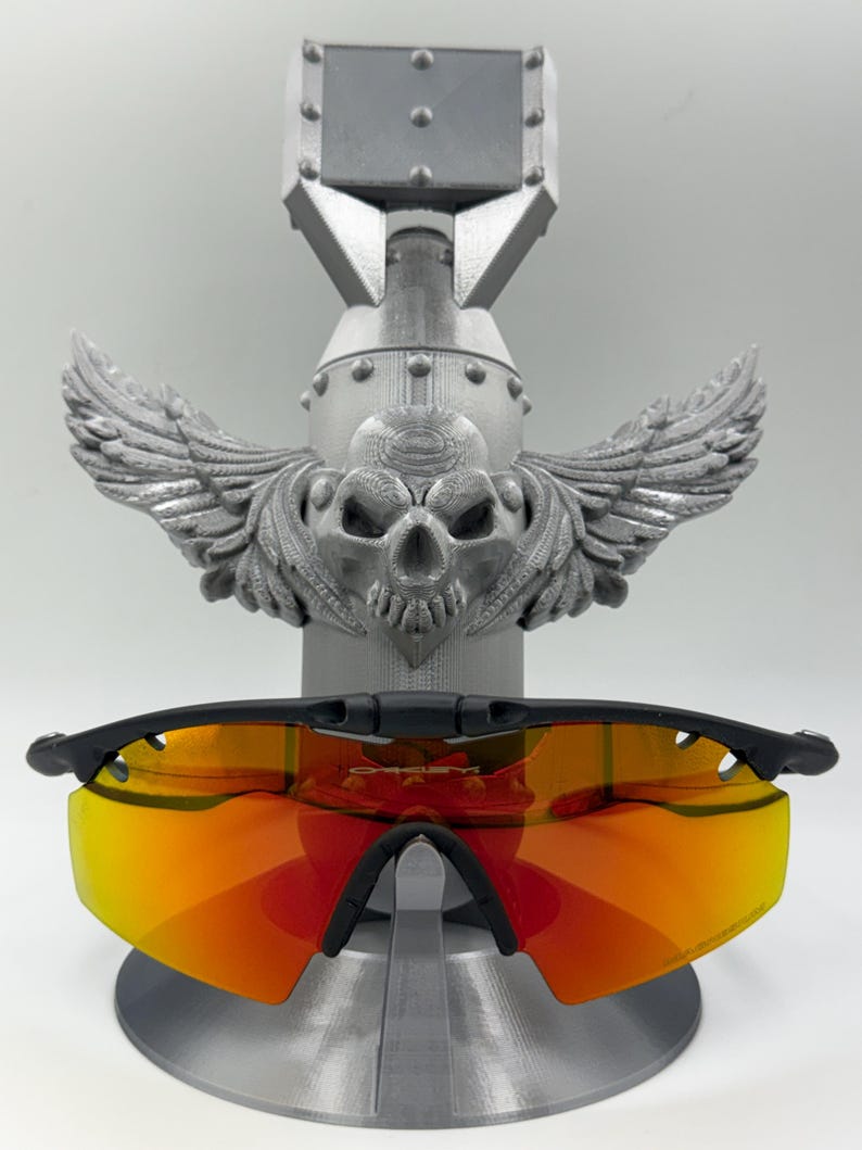 May include: Black-framed sunglasses with orange-red lenses rest on a gray, bomb-shaped display stand. The stand features a skull with wings. The sunglasses are the focal point, showcasing their vibrant lenses and sleek design.