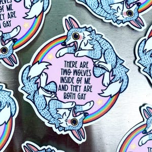 May include: A sticker featuring two cartoon wolves in light blue, set against a pink background with a rainbow and stars. The text reads, "THERE ARE TWO WOLVES INSIDE OF ME AND THEY ARE BOTH GAY."
