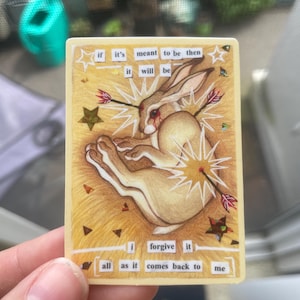 Puede incluir: Una tarjeta rectangular con una ilustración de un conejo atravesado por flechas. La tarjeta tiene el texto "if it's meant to be then it will be" y "i forgive it all as it comes back to me". El fondo es de color marrón claro.