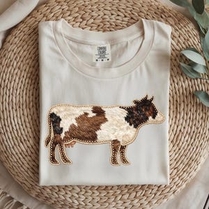 Può includere: T-shirt color crema con un design di mucca ricamato. La mucca ha un motivo marrone, nero e bianco con un contorno dorato. La maglietta è piegata su un tappetino intrecciato.
