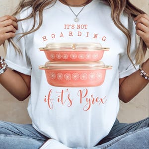 May include: White t-shirt with the text "IT'S NOT HOARDING if it's Pyrex" in coral lettering. Two coral-colored Pyrex dishes with floral patterns are stacked on top of each other. The shirt is being worn by a person.