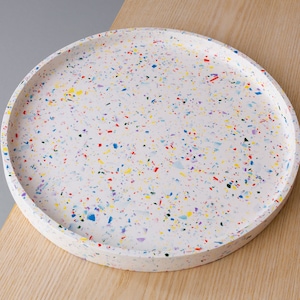 Könnte beinhalten: Ein rundes weißes Tablett mit einem gesprenkelten Muster aus bunten Punkten. Das Tablett besteht aus einem gesprenkelten Material mit roten, gelben, blauen und violetten Punkten.