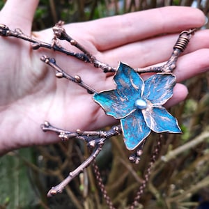 May include: A copper necklace with a blue and green flower pendant and a pearl accent. The necklace is made of twisted copper wire and has a delicate, nature-inspired design.