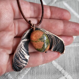 May include: A copper pendant necklace featuring a green, orange, and brown stone set in a copper maple seed design.