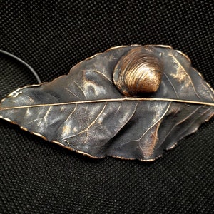 以下が含まれることがあります： 暗いグレーとブロンズの色合いの、詳細な葉のデザインが特徴の手作りのペンダントネックレス。 小さな、質感のあるブロンズ色の貝殻が葉に付いています。 ネックレスは黒いコードが付いています。