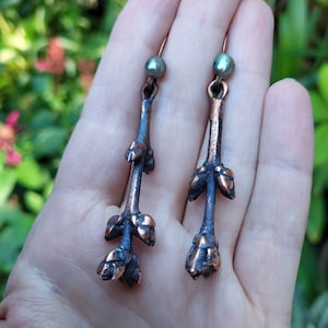 May include: A pair of handcrafted copper earrings. Each earring features a long, slender stem with bud-like details and a small, round, green bead at the top. The earrings have a rustic, natural aesthetic, perfect for everyday wear.