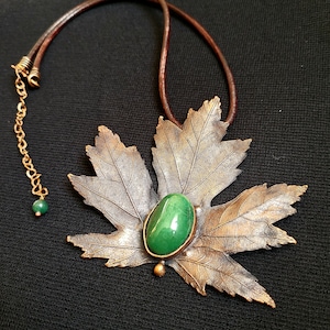 May include: A bronze leaf pendant necklace with a green gemstone in the center. The necklace is on a brown leather cord.