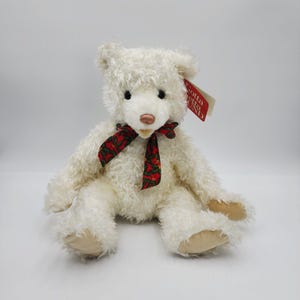 Könnte beinhalten: Ein cremefarbener Plüschteddybär mit zotteligem Fell, einer hellbraunen Nase und beigen Stoffpfoten. Der Bär trägt ein rotes Band mit einem grünen Stechpalmen- und Beerenmuster um den Hals und hat ein rotes Etikett mit der Aufschrift Gotta Getta GUND.