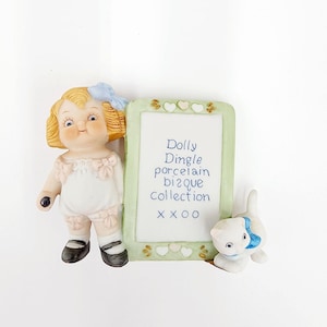 May include: A porcelain figurine of a girl with blonde hair and a blue bow holding a small black object. She is standing next to a green frame with a white background and the text "Dolly Dingle porcelain bisque collection XXXO". A white kitten with a blue bow is standing next to the frame.