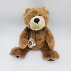 Puede incluir: Un oso de peluche marrón con hocico beige y nariz azul oscuro. El oso tiene una etiqueta que dice "BOYDS". El oso está sentado con las piernas extendidas. El pelaje del oso es suave y esponjoso.