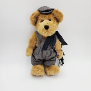 May include: A plush teddy bear with golden-brown fur wearing a dark grey herringbone flat cap and matching overalls with a single button. A dark navy blue patterned ribbon is tied around the neck, and a small tag hangs from the side of the overalls.