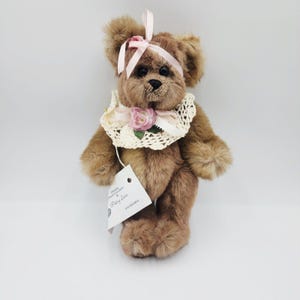 Puede incluir: Un oso de peluche marrón con una cinta rosa en la cabeza y un cuello de encaje. El oso tiene una etiqueta adjunta. Una rosa rosa está unida al cuello. El oso está sobre una superficie blanca.