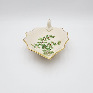 May include: A white ceramic dish with a gold rim and a holly design. The dish has a handle in the shape of a leaf.
