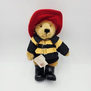 Op de afbeelding: Een pluche teddybeer met lichtbruine vacht verkleed als brandweerman. De beer draagt een felrode vilten helm, een zwart-geel jasje met zilveren gespen en glimmende zwarte laarzen. Een wit papieren label hangt aan de gesp aan de voorzijde van het jasje.