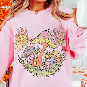 May include: A light pink crew neck sweatshirt with a retro graphic of mushrooms, flowers, a sun, and a butterfly. The design has a whimsical feel. The sweatshirt is a comfortable style.