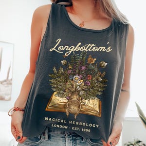 May include: A dark gray tank top with the text "Longbottom's Magical Herbology London - Est. 1996" and an illustration of a book with a plant growing out of it.