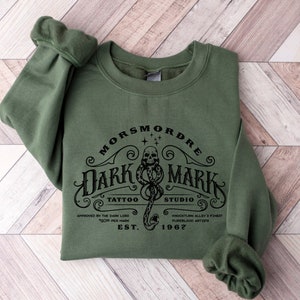 May include: A dark green sweatshirt with a black graphic print of a snake with a skull and the text "MORS MORDE DARK MARK TATTOO STUDIO APPROVED BY THE DARK LORD 100% PURE PER MARK EST. 1967 KNOCHTURN ALLEY'S FINEST PUREBLOOD ARTISTS".
