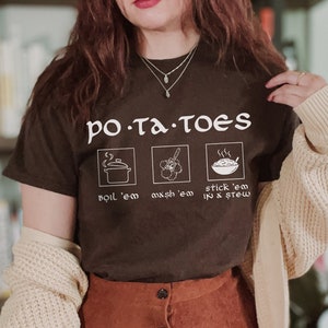 May include: A brown t-shirt with the text "po.ta.toes" and three illustrations of potatoes. The illustrations show a pot of boiling potatoes, mashed potatoes, and potatoes in a stew. The person wearing the shirt is also wearing a brown corduroy skirt and a beige cardigan.