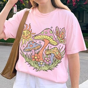 May include: A light pink t-shirt with a vibrant graphic of mushrooms, a sun, and a butterfly. The outfit includes white shorts and a white baseball cap. A brown shoulder bag is also visible.