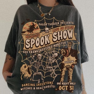 May include: A dark grey t-shirt with a vintage-style graphic print of a spooky Halloween show. The graphic features a witch, skeletons, and Frankenstein's monster. The text reads "Salem Theatre Presents Midnight Spook Show Live on Stage See Frankenstein's Monster In Person! Dancing Skeletons Witches & Real Ghosts! One Night Only Oct 31."