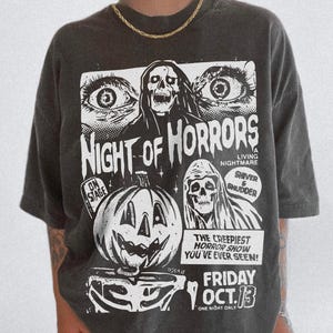 May include: Dark grey t-shirt with a Halloween graphic. The design features the text "Night of Horrors", a skull, a jack-o'-lantern, and the text "Friday Oct. 13". The shirt has short sleeves and a loose fit. A spooky design for the Halloween season.