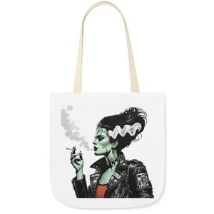 May include: A white tote bag with a black and white illustration of a green-skinned woman with stitched scars on her face and neck. She is wearing a black leather jacket with studs and a red shirt. She is smoking a cigarette and has a hairstyle with a wave in the front.