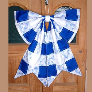 May include: A large, decorative bow made of fabric, featuring a patchwork design of white fabric with blue floral patterns and solid blue squares. The bow is suspended by a white cord, suitable for door or wall decor.