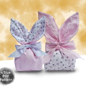 May include: Two pink fabric bunny-shaped gift bags with large, floppy ears. One has a white bow, the other a pink bow. The ears are lined with a floral pattern. The bags are tied at the base with a bow.