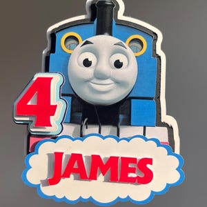 Könnte beinhalten: Ein blau-weißer Tortenaufsatz mit Thomas der kleinen Lokomotive, der die rote Zahl 4 und den Namen "JAMES" in roten Buchstaben auf einem wolkenförmigen Banner zeigt. Für eine Geburtstagsfeier.