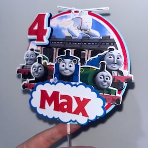 Könnte beinhalten: Ein farbenfroher Tortenaufsatz mit der Zahl 4 und Abbildungen von Zeichentrickfiguren von Zügen. Der Name "Max" ist auf einem wolkenförmigen Banner zu sehen. Ein Hubschrauber ist ebenfalls sichtbar. Der Aufsatz wird von einem transparenten Kunststoffstab gehalten.