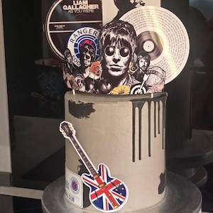 以下が含まれることがあります： 音楽をテーマにした画像とユニオンジャックのデザインのギターのステッカーで飾られた灰色のケーキ。ケーキにはミュージシャンとレコードの画像があり、黒いアイシングが垂れています。