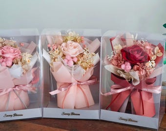 May include: Three clear boxes each containing a small bouquet of preserved flowers. The bouquets are wrapped in pink, red, and white tissue paper and tied with a ribbon. The boxes are labeled "Sunny Blossom".