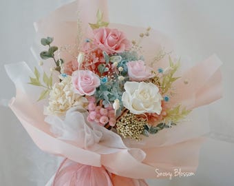 May include: A bouquet of preserved flowers featuring pink, white, and light blue roses, hydrangea, and small filler plants. The arrangement is wrapped in layered pale pink paper and tied with a matching pink ribbon. The text Snowy Blossom is visible.
