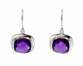 Op de afbeelding: Een paar zilveren oorbellen met vierkant geslepen, gefacetteerde paarse amethist edelstenen in gepolijste zilveren zettingen. Elke oorbel hangt aan een eenvoudige zilveren haak en toont de diepe violette kleur en geometrische helderheid.