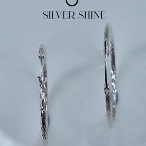 Op de afbeelding: Twee zilveren oorringen met een textuur. De oorbellen worden gepresenteerd tegen een lichte achtergrond, met de tekst "SILVER SHINE" erboven.