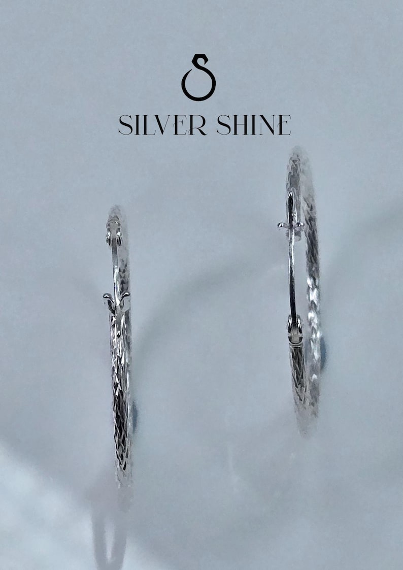 Op de afbeelding: Twee zilveren oorringen met een textuur. De oorbellen worden gepresenteerd tegen een lichte achtergrond, met de tekst "SILVER SHINE" erboven.