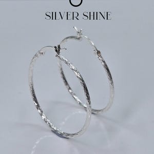 Op de afbeelding: Een paar zilveren oorringen met een getextureerd oppervlak. De oorbellen worden op een witte achtergrond weergegeven. De tekst "SILVER SHINE" staat boven de oorbellen.