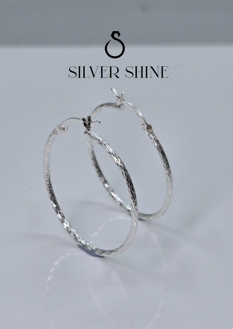 Op de afbeelding: Een paar zilveren oorringen met een getextureerd oppervlak. De oorbellen worden op een witte achtergrond weergegeven. De tekst "SILVER SHINE" staat boven de oorbellen.