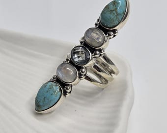 May include: A long, silver ring featuring five stones. Two teardrop-shaped turquoise stones flank three smaller stones: two round, iridescent moonstones and a square, clear stone. The ring has a multi-band design.