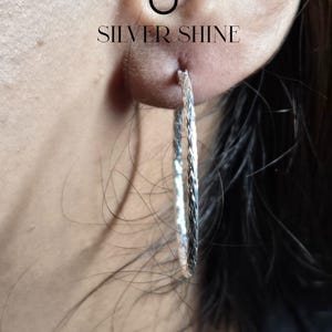 Op de afbeelding: Close-up van een zilveren oorring met een textuurontwerp. De oorring wordt gedragen in een oor piercing. De tekst "SILVER SHINE" is zichtbaar boven het oor.