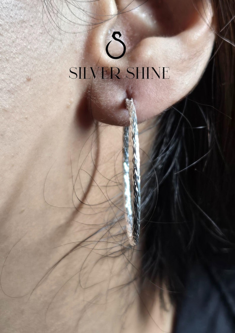 Op de afbeelding: Close-up van een zilveren oorring met een textuurontwerp. De oorring wordt gedragen in een oor piercing. De tekst "SILVER SHINE" is zichtbaar boven het oor.