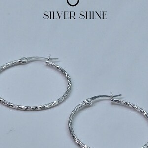 Op de afbeelding: Een paar zilveren oorringen met een getextureerd, diamantgeslepen patroon. De oorbellen hebben een scharniersluiting en worden tegen een witte achtergrond getoond. De tekst "SILVER SHINE" staat erboven.