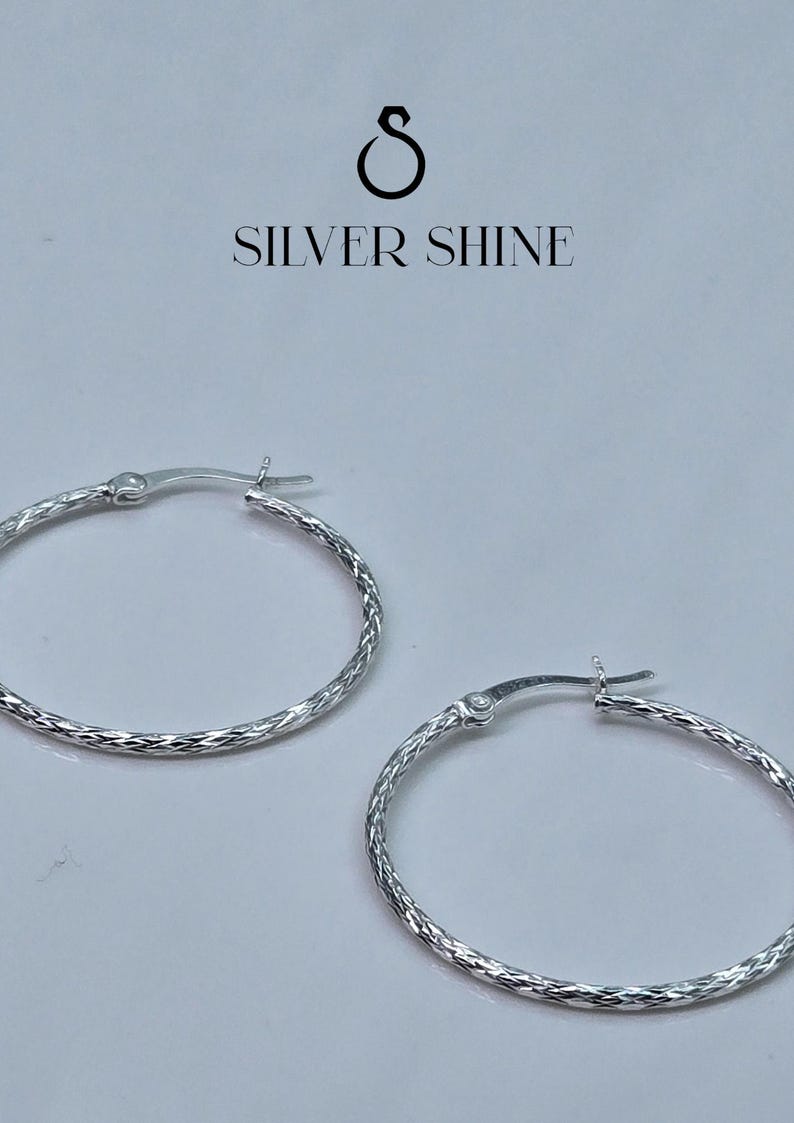 Op de afbeelding: Een paar zilveren oorringen met een getextureerd, diamantgeslepen patroon. De oorbellen hebben een scharniersluiting en worden tegen een witte achtergrond getoond. De tekst "SILVER SHINE" staat erboven.