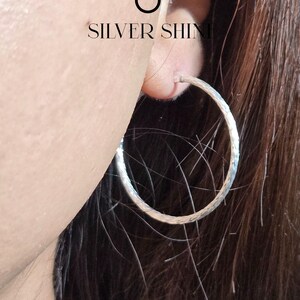 Op de afbeelding: Close-up van een zilveren oorring met een getextureerd oppervlak. De oorring wordt in een oor gedragen, met de tekst "SILVER SHINE" en een zichtbaar logo. De oorring heeft een cirkelvorm.
