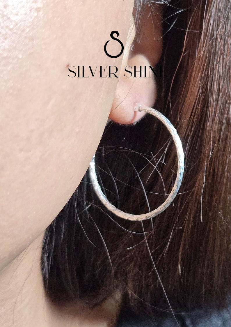 Op de afbeelding: Close-up van een zilveren oorring met een getextureerd oppervlak. De oorring wordt in een oor gedragen, met de tekst "SILVER SHINE" en een zichtbaar logo. De oorring heeft een cirkelvorm.