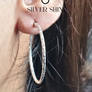 Op de afbeelding: Close-up van een zilveren oorring met een textuurontwerp. De oorring wordt op een oor gedragen, met de tekst "SILVER SHINE" zichtbaar boven de oorring. Een klassiek sieraad.