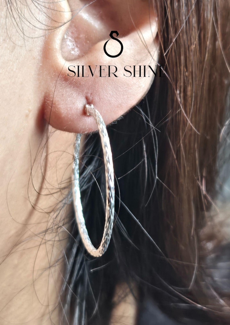 Op de afbeelding: Close-up van een zilveren oorring met een textuurontwerp. De oorring wordt op een oor gedragen, met de tekst "SILVER SHINE" zichtbaar boven de oorring. Een klassiek sieraad.