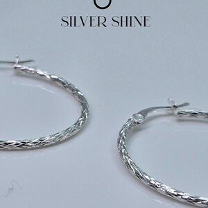 Op de afbeelding: Close-up van twee zilveren oorringen met een getextureerd, diamantgeslepen patroon. De oorbellen worden gepresenteerd tegen een witte achtergrond. De tekst "SILVER SHINE" staat erboven.
