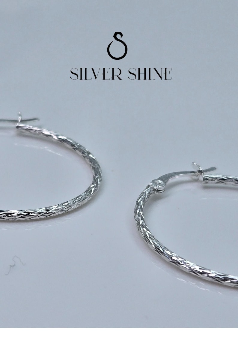 Op de afbeelding: Close-up van twee zilveren oorringen met een getextureerd, diamantgeslepen patroon. De oorbellen worden gepresenteerd tegen een witte achtergrond. De tekst "SILVER SHINE" staat erboven.