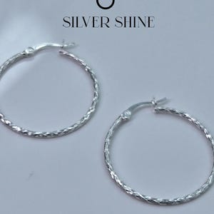 Op de afbeelding: Twee zilveren oorringen met een getextureerd, gedraaid ontwerp. De oorbellen worden gepresenteerd op een lichtgrijze achtergrond. De tekst "SILVER SHINE" staat boven de oorbellen.